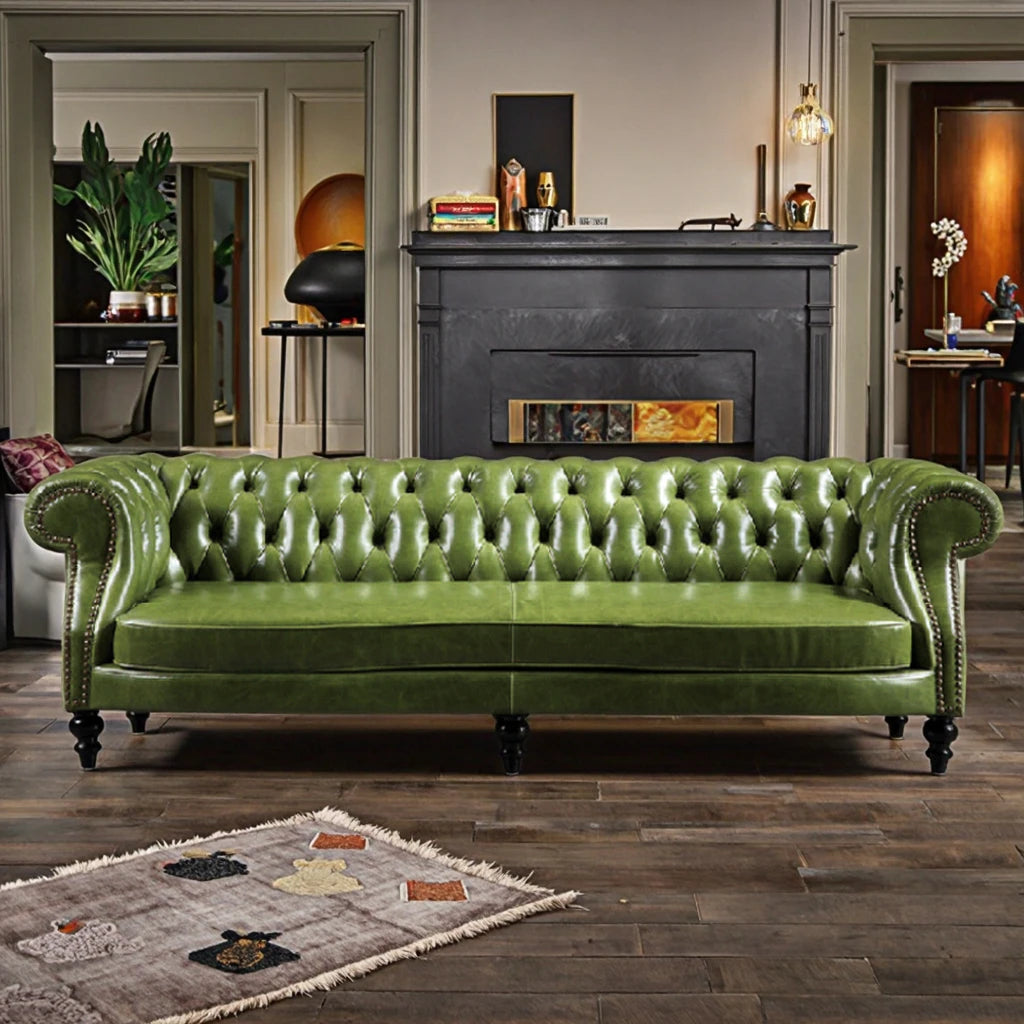A green tufted leather Chesterfield sofa with nailhead trim and dark wooden legs, positioned in front of a black fireplace in a styled living room.