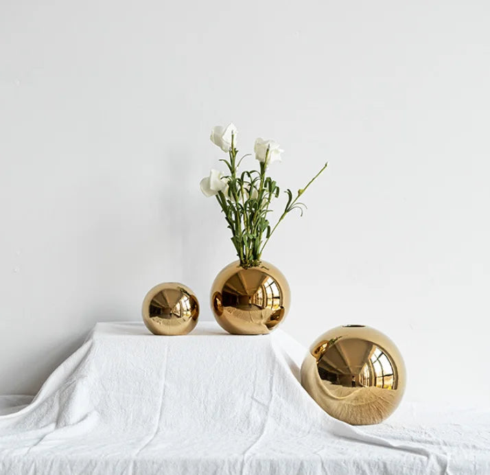 Three round metallic gold vases of different sizes arranged on a white draped surface against a white wall, with white flowers placed in the center vase.