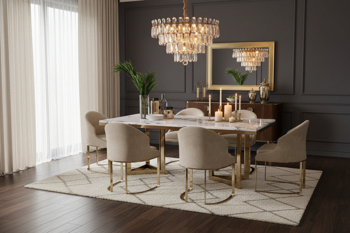 A modern dining table set featuring a rectangular white marble tabletop with a gold metal frame, surrounded by six upholstered dining chairs in beige velvet with matching gold metal legs. The set is styled with a vase of green palm leaves and small tabletop accessories on dark background.  Chandelier overhead and a decorative mirror