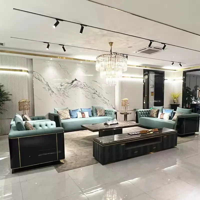 A luxury modern living room set featuring a teal tufted sofa, a matching armchair, and a dark wood coffee table with gold trim, arranged in a high-ceilinged showroom with a crystal chandelier and marble wall.