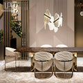 A modern dining room setup featuring a dark wood rectangular table with a polished gold metal base, surrounded by four white marble-patterned upholstered dining chairs with gold metal frames. A unique multi-arm pendant light hangs above the table against a geometric beige wall.