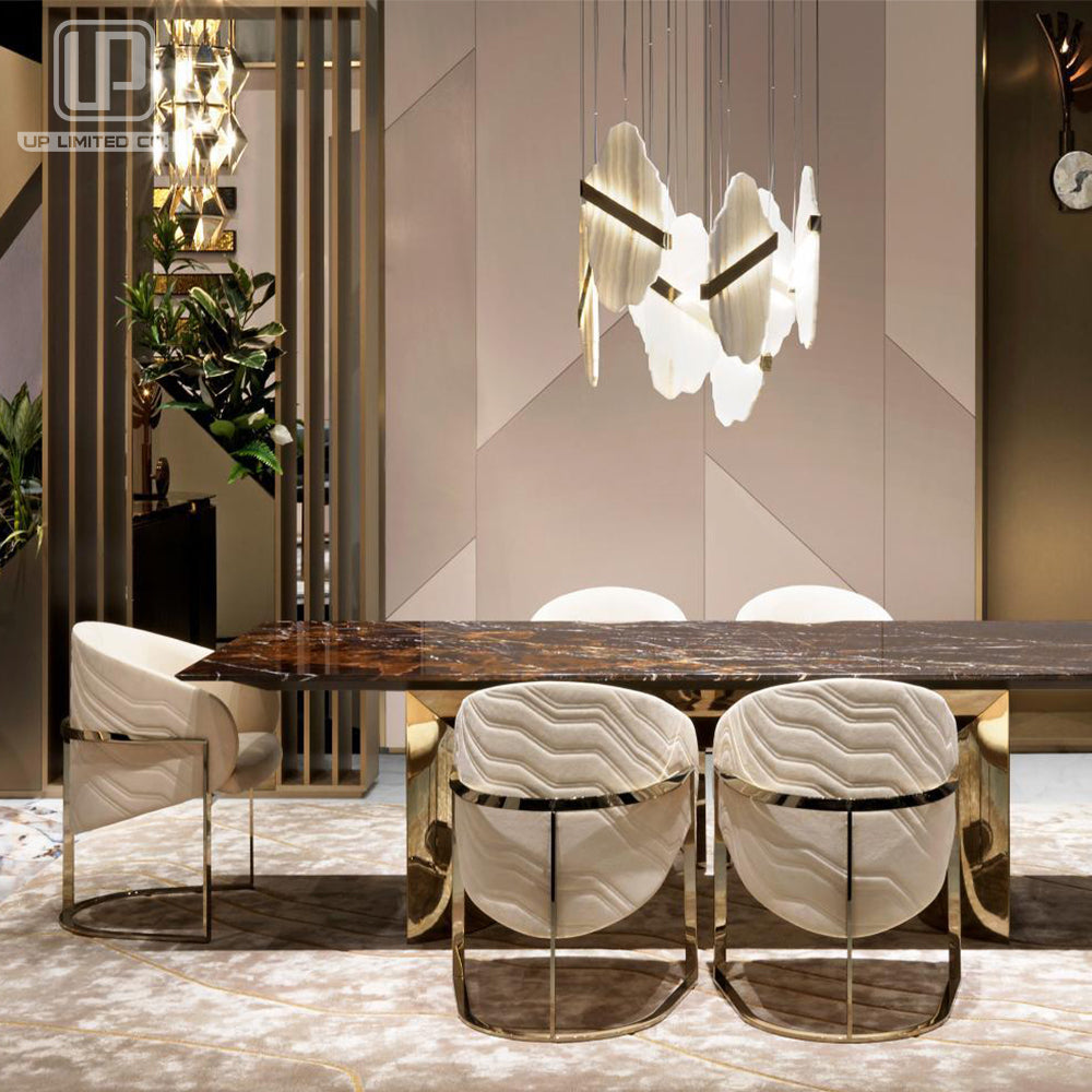A modern dining room setup featuring a dark wood rectangular table with a polished gold metal base, surrounded by four white marble-patterned upholstered dining chairs with gold metal frames. A unique multi-arm pendant light hangs above the table against a geometric beige wall.