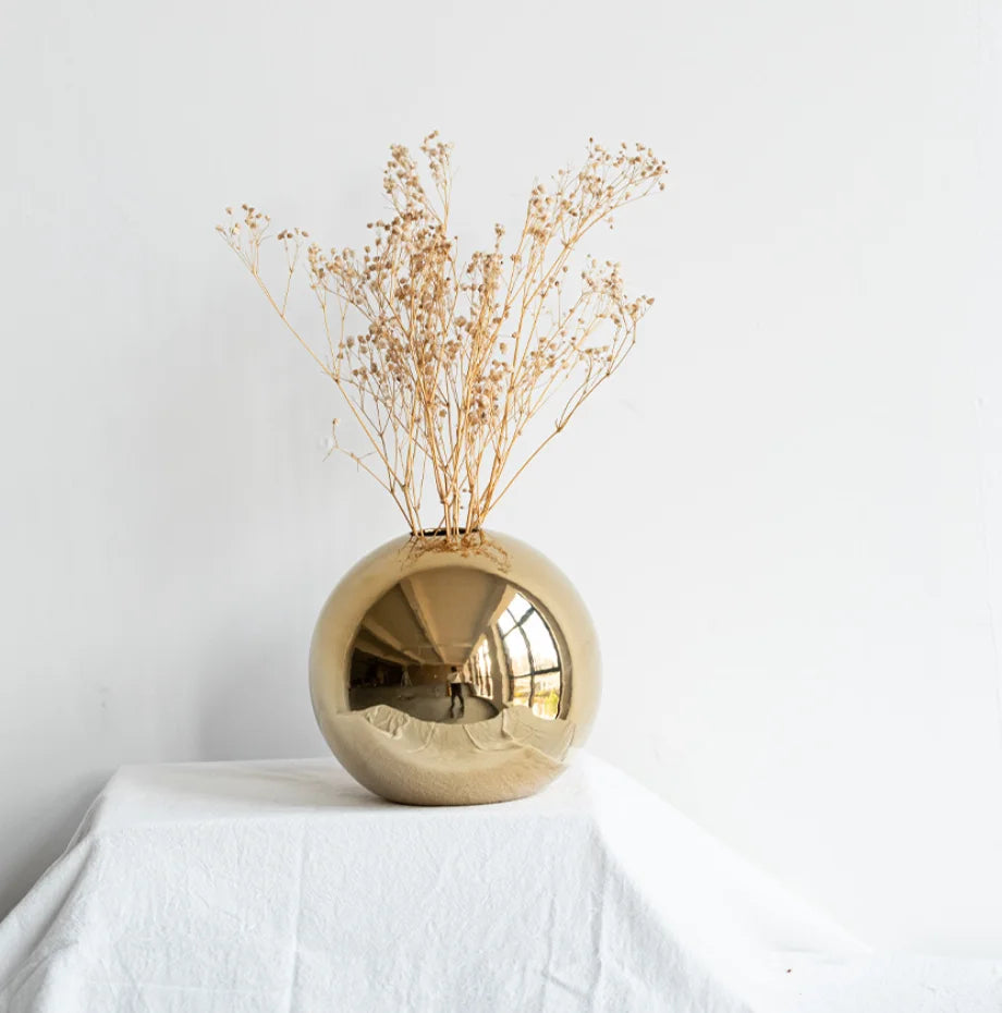 A shiny spherical gold vase filled with dried beige flowers, displayed on a white draped surface against a white wall.