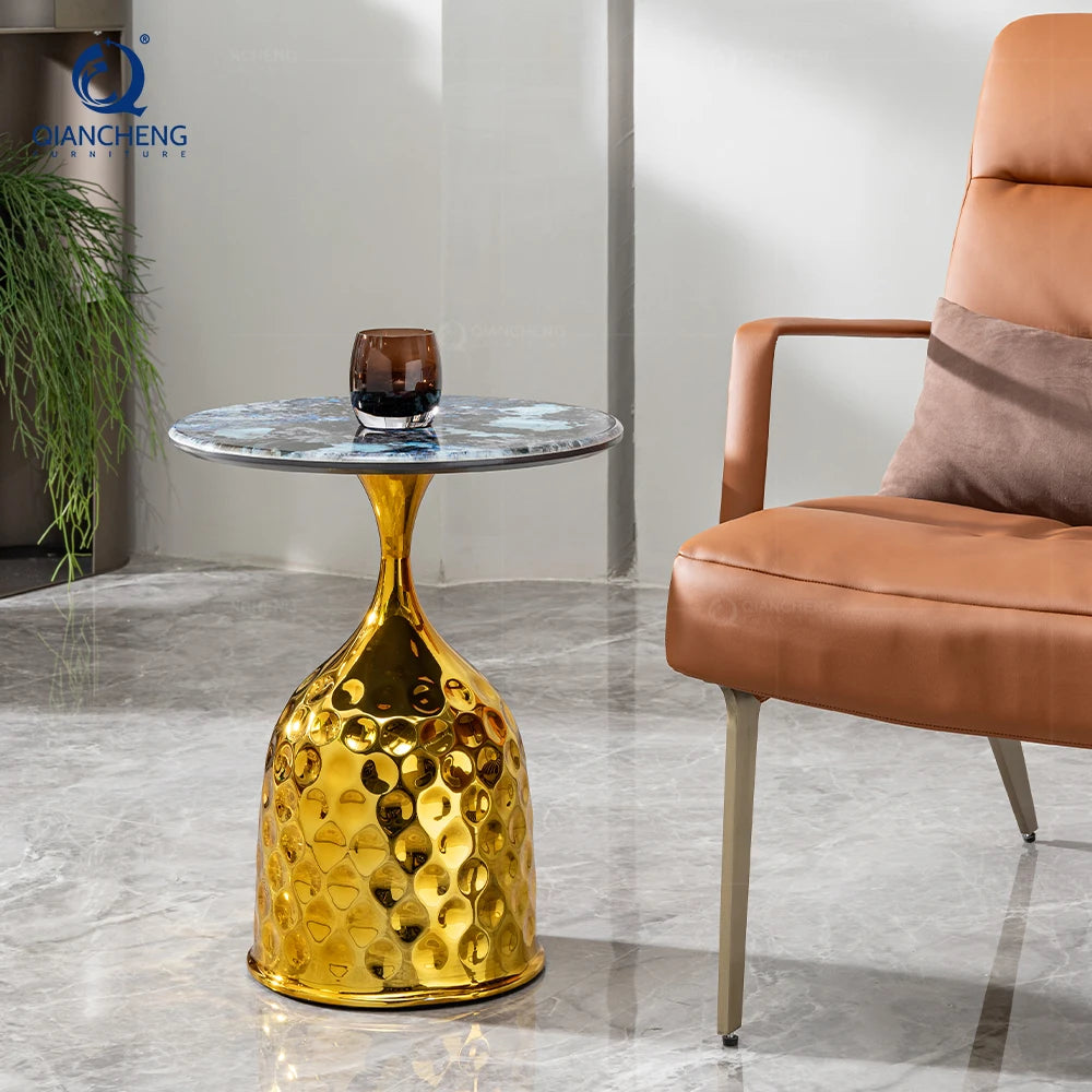 Gold side table with a glass on a marble floor next to a brown chair.