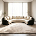 A modern curved sectional sofa with cream tufted cushions and black leather arms, placed on a large beige rug in a bright minimalist room with tall windows and sheer curtains.