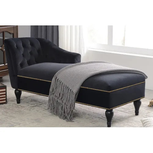 A black velvet chaise lounge with gold nailhead trim and dark wooden legs, styled with a gray throw blanket in a bright room.