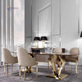 A modern dining room setup featuring a rectangular glass-top table with a geometric gold metal base, surrounded by four upholstered dining chairs in beige and cream tones with gold metal legs. The background consists of sleek, light gray paneled walls and large windows.