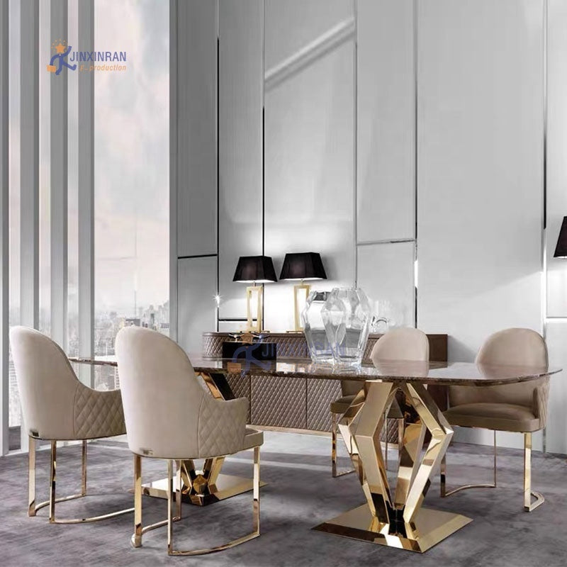 A modern dining room setup featuring a rectangular glass-top table with a geometric gold metal base, surrounded by four upholstered dining chairs in beige and cream tones with gold metal legs. The background consists of sleek, light gray paneled walls and large windows.