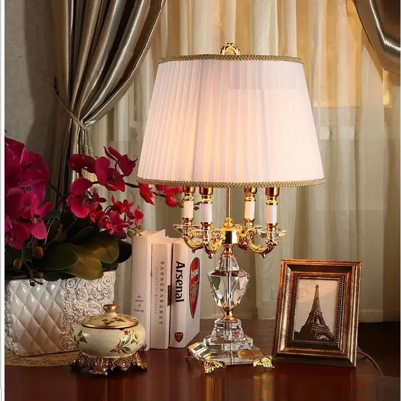K9 crystal table lamp with fabric shade on a side table with flowers, creating a warm ambiance in a stylish room.