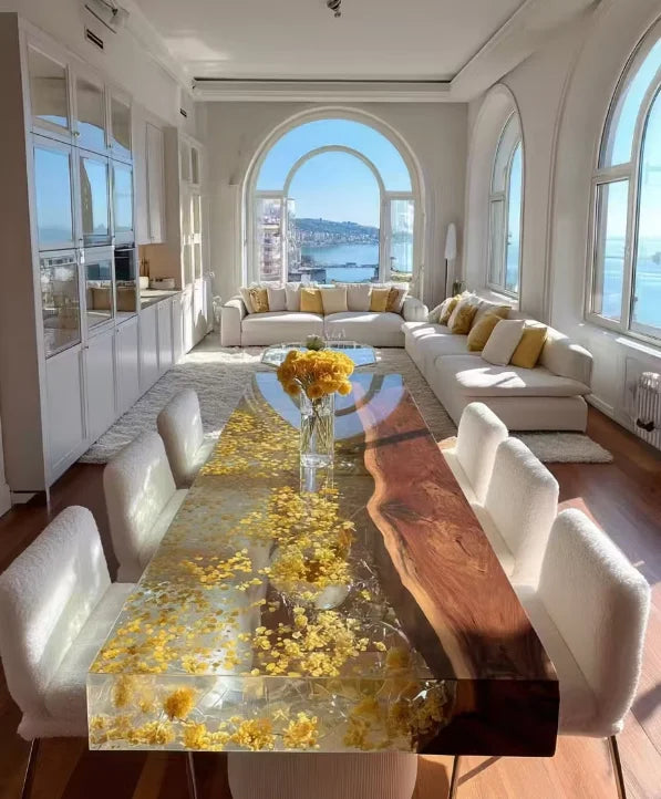 A long dining table with a live-edge natural wood slab and a clear resin top filled with yellow flower petals, surrounded by white upholstered chairs in a bright, sunlit modern dining room with large arched windows overlooking a waterfront.