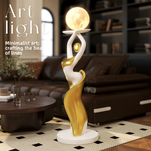 A modern abstract sculpture lamp in a living room setting, featuring a white and gold female figure holding a glowing moon-like orb on her head. The sculpture stands on a round white base and is illuminated, set against a blurred background of a brown leather sofa and bookshelves.