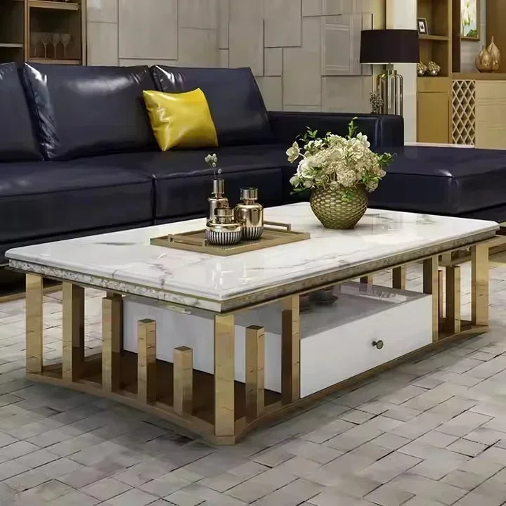 A modern rectangular coffee table with a white marble-patterned top and a light wood frame, featuring an open lower drawer and decorative side supports. The table is styled in a living room setting with a dark leather sofa and a vase of flowers on top.