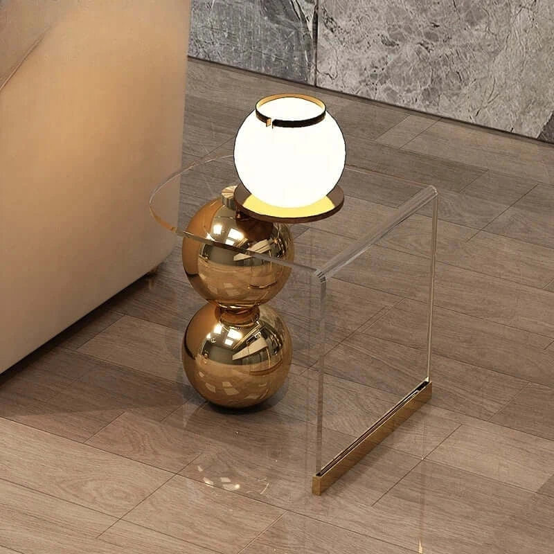 Modern gold side table with a clear glass shelf and white lamp on a wooden floor.