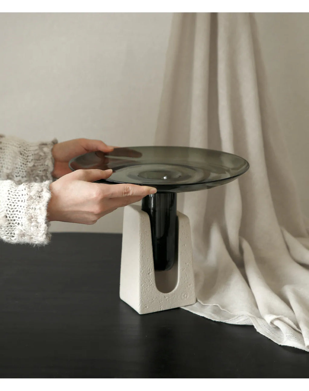 A modern pedestal table with a square concrete-look base and a dark smoked glass top, held by a person wearing a white knit sweater against a neutral draped backdrop.