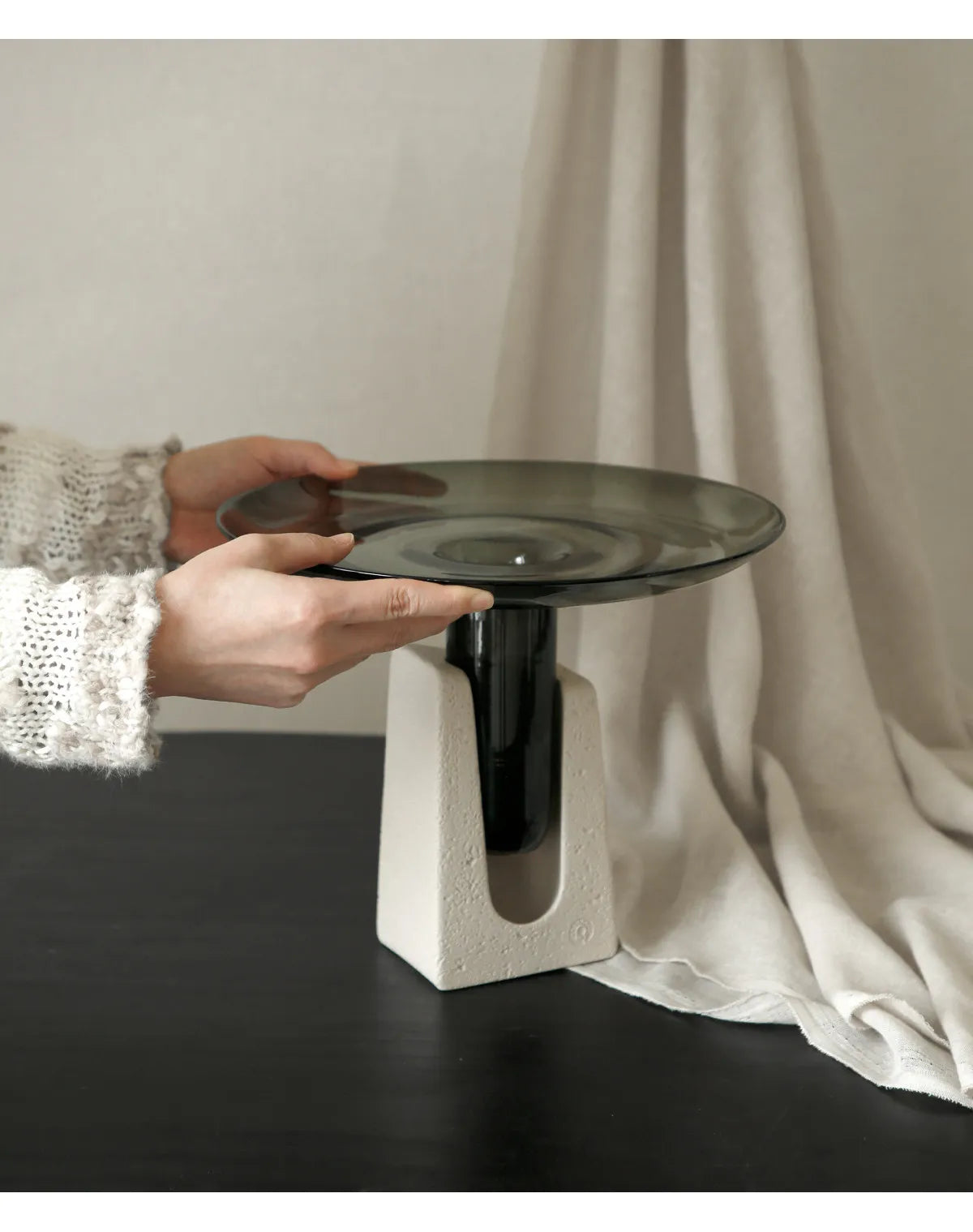 A modern pedestal table with a square concrete-look base and a dark smoked glass top, held by a person wearing a white knit sweater against a neutral draped backdrop.