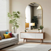 Arched metal framed bathroom mirror hanging above modern console table with plants and decorative items in bright room.