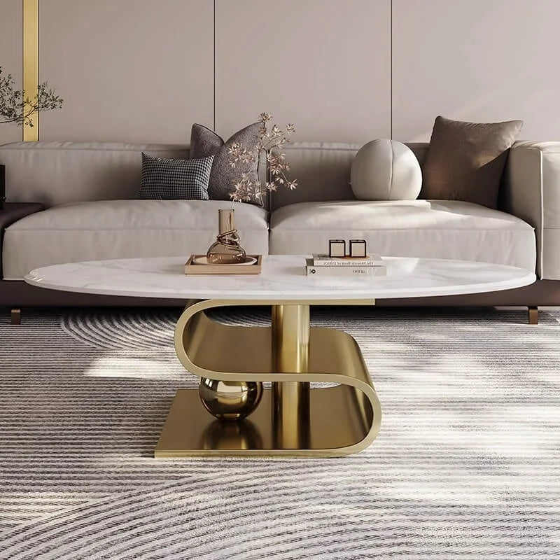 A modern oval coffee table with a white marble-look top and a sculptural curved gold metal base, styled in a contemporary living room in front of a light beige sofa.