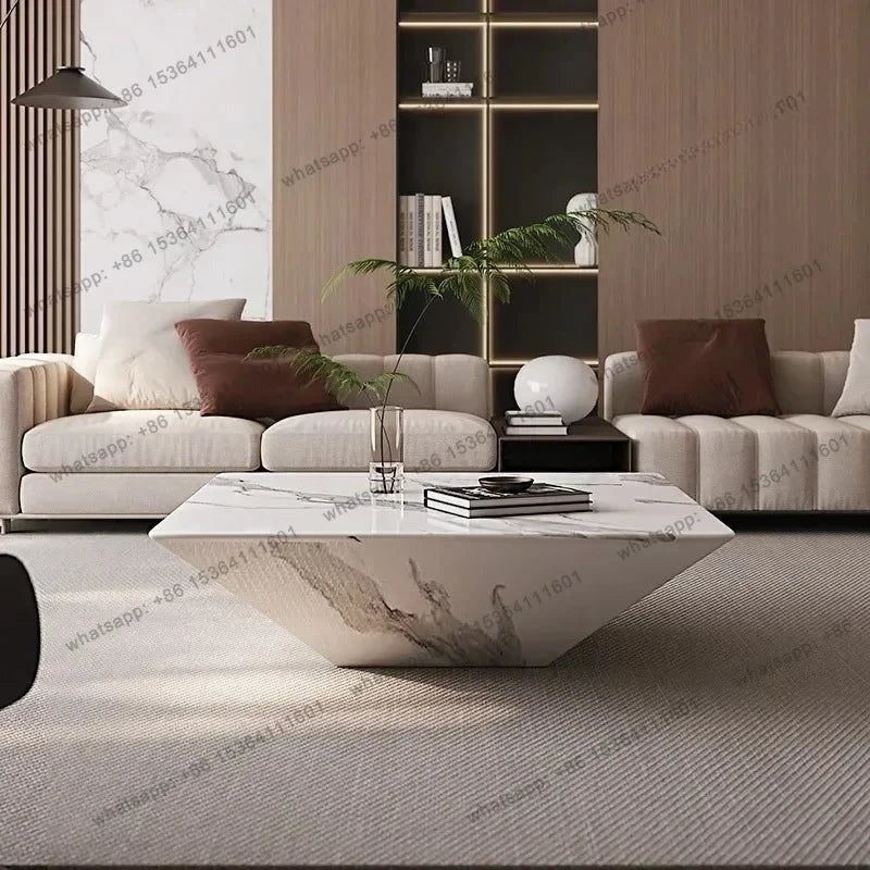 A modern living room featuring a geometric white marble coffee table with gray veining, styled with a vase of greenery and books. In the background are two cream-colored sofas with brown cushions, a dark wood shelving unit, and a textured light gray rug.