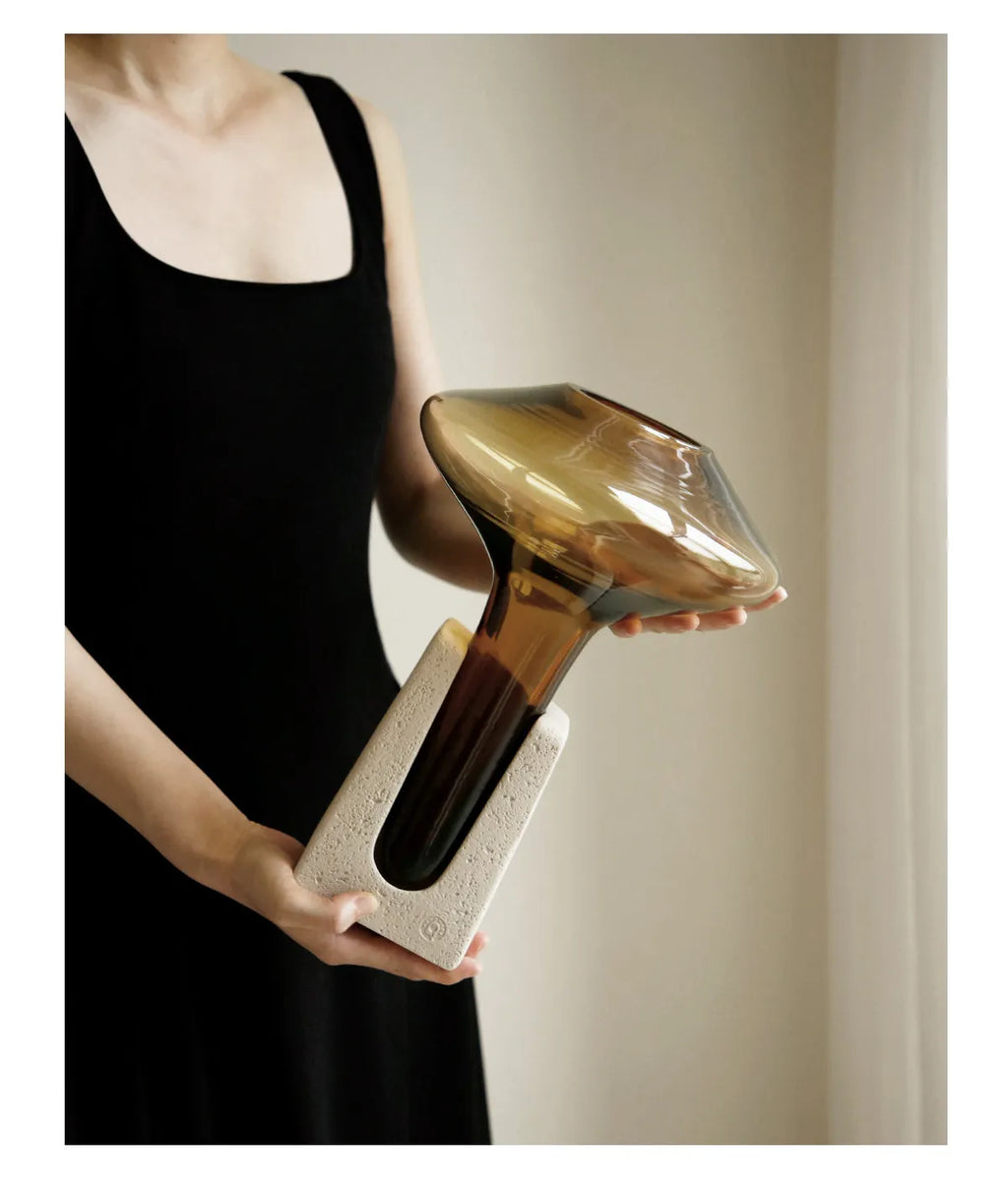 A person in a black sleeveless dress holds a sculptural amber-brown glass vessel with a wide rounded top and a textured off-white base against a soft beige indoor background.