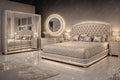 A luxury bedroom set featuring a bed with a tufted, silver-embellished headboard and matching silver nightstands, a large mirrored wardrobe with silver trim, and a glowing round wall mirror against a dark gray wall.