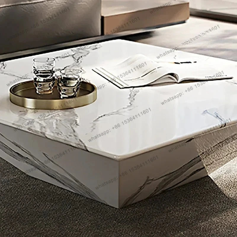 A square coffee table with a white and gray marble pattern, styled with a gold tray, two glasses, and an open magazine. The table is set in a modern living room environment.