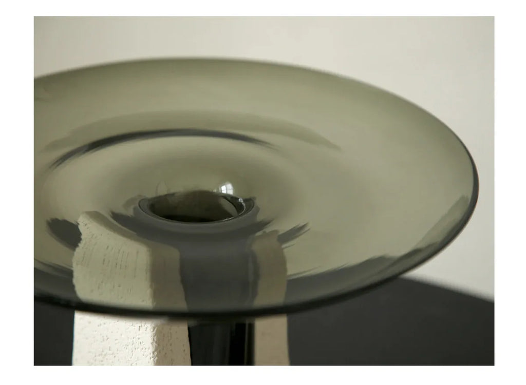 A close-up of a translucent smoky gray glass table top with a circular center opening, supported by a textured off-white pedestal against a neutral light background.