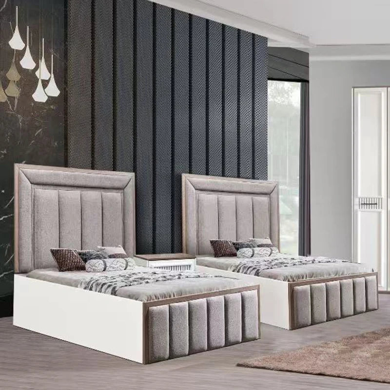 Two matching single beds with tall upholstered headboards and footboards in a modern bedroom. The beds have white storage bases, light wood trim, and gray upholstered panels, styled with gray and brown patterned bedding and pillows.