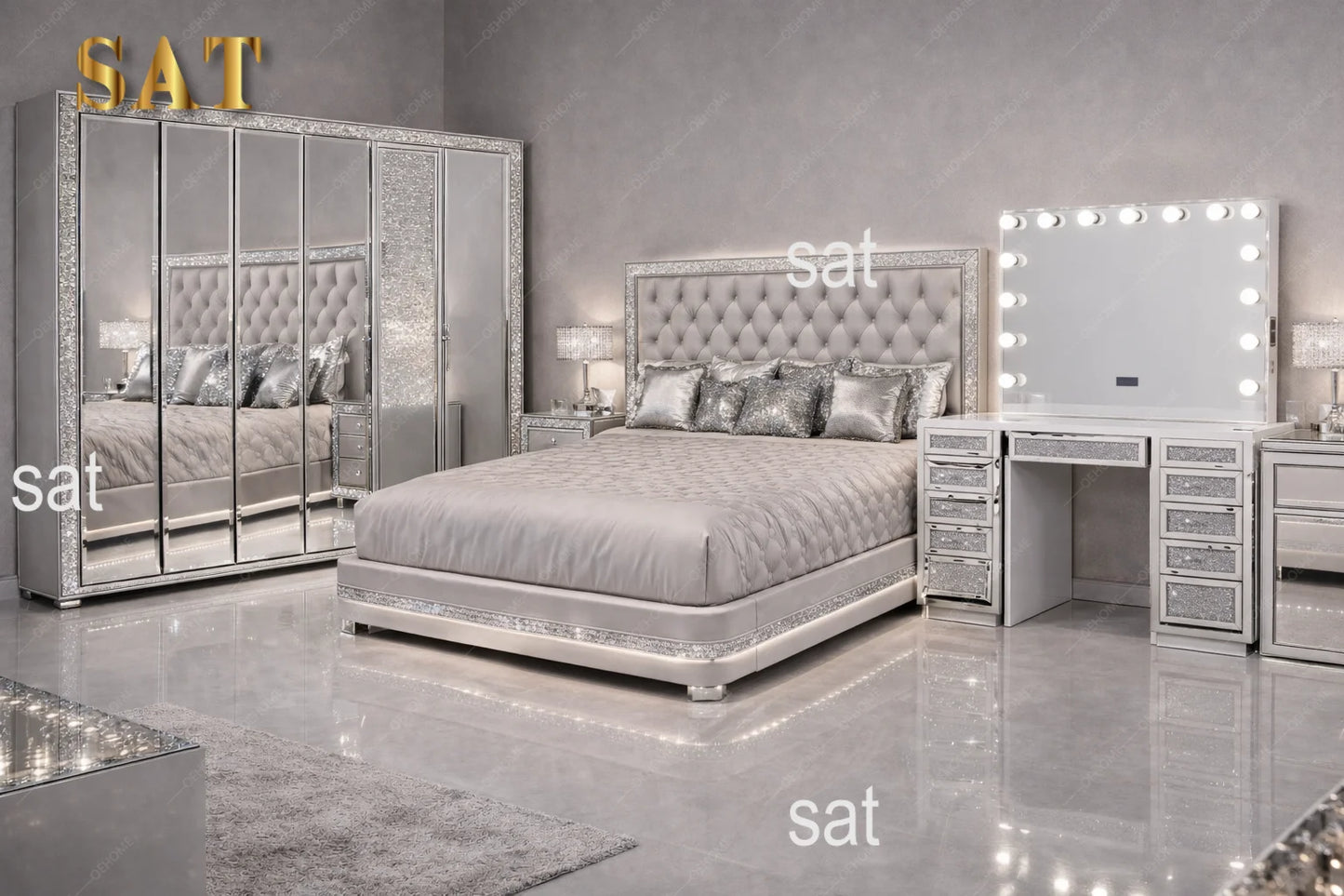A luxury bedroom set featuring a silver tufted bed frame, a matching mirrored wardrobe with gold 'SAT' lettering, a vanity table with drawers, and a lighted makeup mirror, all in a high-gloss silver finish.