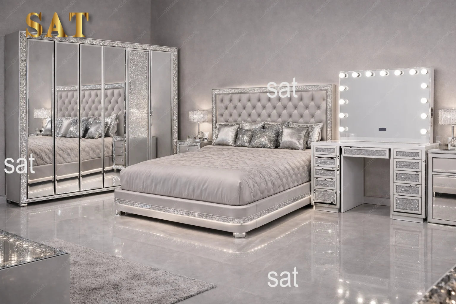 A luxury bedroom set featuring a silver tufted bed frame, a matching mirrored wardrobe with gold 'SAT' lettering, a vanity table with drawers, and a lighted makeup mirror, all in a high-gloss silver finish.