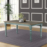 Wooden dining table with teal legs in a room with gray walls and large windows.